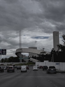 A busy road with multiple cars heading towards a prominent structure that includes a large archway labeled 'Tecnol&oacute;gico de Monterrey'. The background shows a tall building with a distinct logo near the top, surrounded by trees and greenery. The sky above is overcast with thick clouds, contributing to a somewhat gloomy atmosphere.