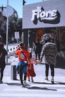 Pedestrians walk along a sidewalk near a store with a 'Fiore' sign. The scene includes adults and children, wearing casual clothing, and is set in an urban area with traffic signs in the background.