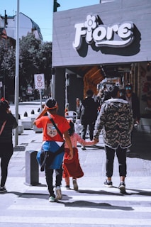 Pedestrians walk along a sidewalk near a store with a 'Fiore' sign. The scene includes adults and children, wearing casual clothing, and is set in an urban area with traffic signs in the background.