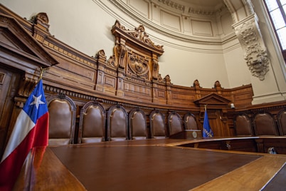 A grandiose courtroom with intricately carved wooden paneling and high ceilings. A row of tall, brown leather chairs lines the back, situated beneath an elaborate decorative relief. The room features a large wooden desk in the foreground. Two flags are present, one with a star on a blue background and red and white stripes, and the other with blue and yellow accents.