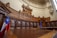 A grandiose courtroom with intricately carved wooden paneling and high ceilings. A row of tall, brown leather chairs lines the back, situated beneath an elaborate decorative relief. The room features a large wooden desk in the foreground. Two flags are present, one with a star on a blue background and red and white stripes, and the other with blue and yellow accents.