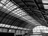 An intricate and expansive architectural structure made of metal and glass dominates the scene. The arched ceiling features a series of repeating patterns formed by iron beams and large translucent panels, allowing light to filter through. The design exemplifies industrial elegance, with a combination of curves and straight lines.