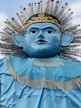A large, stylized blue face with exaggerated features including big ears and bold eyebrows. It has a golden crown with intricate designs on top and a wide cape adorned with golden fringe. The figure is framed by a backdrop of radiating elements resembling a headdress.