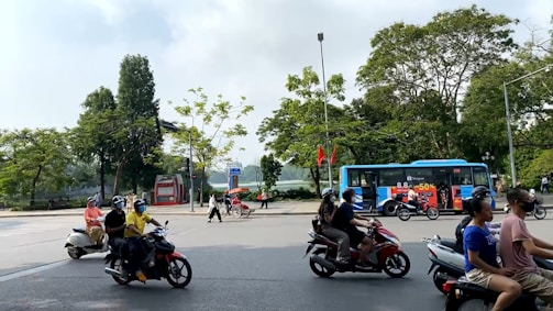A busy street scene features multiple motorcyclists riding past a public bus adorned with advertisements. Trees with green foliage line the street, and several pedestrians are crossing or walking along the sidewalks. Bright clothing and helmets are visible on the motorcycle riders.