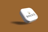 A white, three-dimensional, rectangular object with rounded edges is prominently displayed against a brown background. The object features the word 'Rolex' and the brand's iconic crown logo, both in black, with the crown above the text.