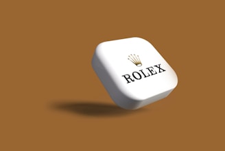 A white, three-dimensional, rectangular object with rounded edges is prominently displayed against a brown background. The object features the word 'Rolex' and the brand's iconic crown logo, both in black, with the crown above the text.