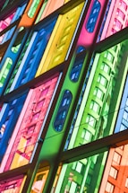 A vibrant, multi-colored glass facade with a geometric design pattern. The glass panels create reflections of a building's curved architecture in a variety of colors, including blue, green, yellow, and pink.
