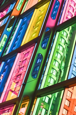A vibrant, multi-colored glass facade with a geometric design pattern. The glass panels create reflections of a building's curved architecture in a variety of colors, including blue, green, yellow, and pink.