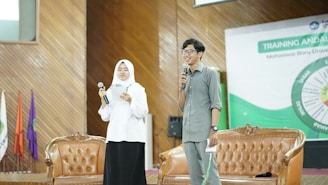 Two individuals stand on a stage holding microphones. One is wearing a white hijab and is holding a piece of paper, while the other is wearing glasses and a gray shirt, smiling as they speak. They are standing in front of two brown leather chairs with a banner in the background that suggests some kind of training or educational event.