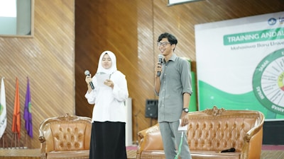 Two individuals stand on a stage holding microphones. One is wearing a white hijab and is holding a piece of paper, while the other is wearing glasses and a gray shirt, smiling as they speak. They are standing in front of two brown leather chairs with a banner in the background that suggests some kind of training or educational event.