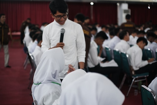 A group of people dressed in formal white shirts are seated in a large room with red carpeting and dark red curtains. A person standing and holding a microphone is interacting with another individual dressed in white, likely responding to a question.