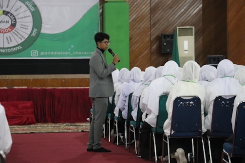 A man in a gray suit is standing and speaking into a microphone in front of a group of seated people wearing white attire. The room has a large circular chart on the wall with green and red sections, and there's a wooden texture on the back wall. The audience is seated in rows of blue chairs.