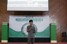 A person is standing on a stage holding a microphone, speaking in front of a large banner. The setting appears to be a formal training or seminar event. The banner contains text about 'Training Andalasian Character' and shows some logos, emphasizing educational or character-building themes. The decor includes wooden paneling and green and white elements.
