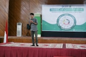 A young person stands on a stage holding a microphone. The backdrop features banners with text about character training, and a diagram with various values highlighted. The stage is covered with a patterned red carpet, and there's an Indonesian flag to one side.