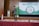 A young person stands on a stage holding a microphone. The backdrop features banners with text about character training, and a diagram with various values highlighted. The stage is covered with a patterned red carpet, and there's an Indonesian flag to one side.