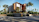 A modern, angular house with a sleek design, featuring a mix of glass, wood, and concrete materials. It is surrounded by tall palm trees and lush greenery, located at the corner of a street with a sports car parked nearby.