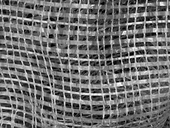 A close-up view of a woven mesh material creates a grid-like pattern. The texture is intricate, with intersecting lines forming a series of squares. The material appears translucent and shiny, giving it a reflective quality.