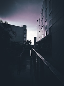 Urban architecture with tall buildings on either side of a pathway, captured under a cloudy sky. The scene is shadowy, enhanced by the moody lighting and reflections on the wet surface.