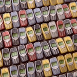 An array of vintage mobile phones, each with a small monochrome screen and physical keypad, is arranged in a pattern. The phones are displayed in multiple colors including red, blue, yellow, and gray, showcasing a nostalgic design typical of early mobile devices.