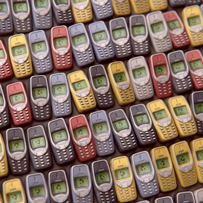 An array of vintage mobile phones, each with a small monochrome screen and physical keypad, is arranged in a pattern. The phones are displayed in multiple colors including red, blue, yellow, and gray, showcasing a nostalgic design typical of early mobile devices.