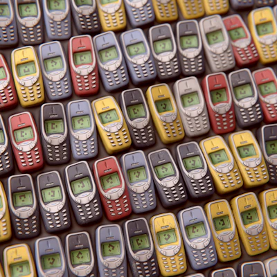 An array of vintage mobile phones, each with a small monochrome screen and physical keypad, is arranged in a pattern. The phones are displayed in multiple colors including red, blue, yellow, and gray, showcasing a nostalgic design typical of early mobile devices.