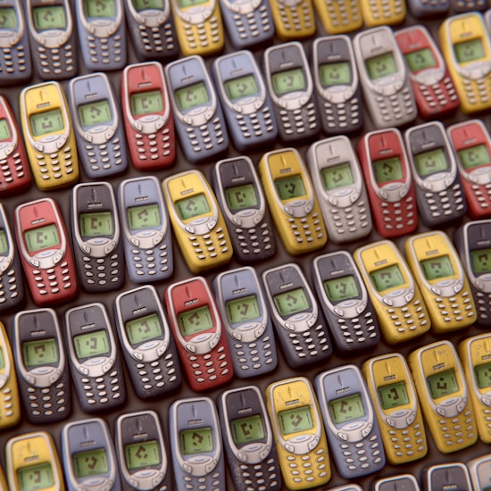 An array of vintage mobile phones, each with a small monochrome screen and physical keypad, is arranged in a pattern. The phones are displayed in multiple colors including red, blue, yellow, and gray, showcasing a nostalgic design typical of early mobile devices.