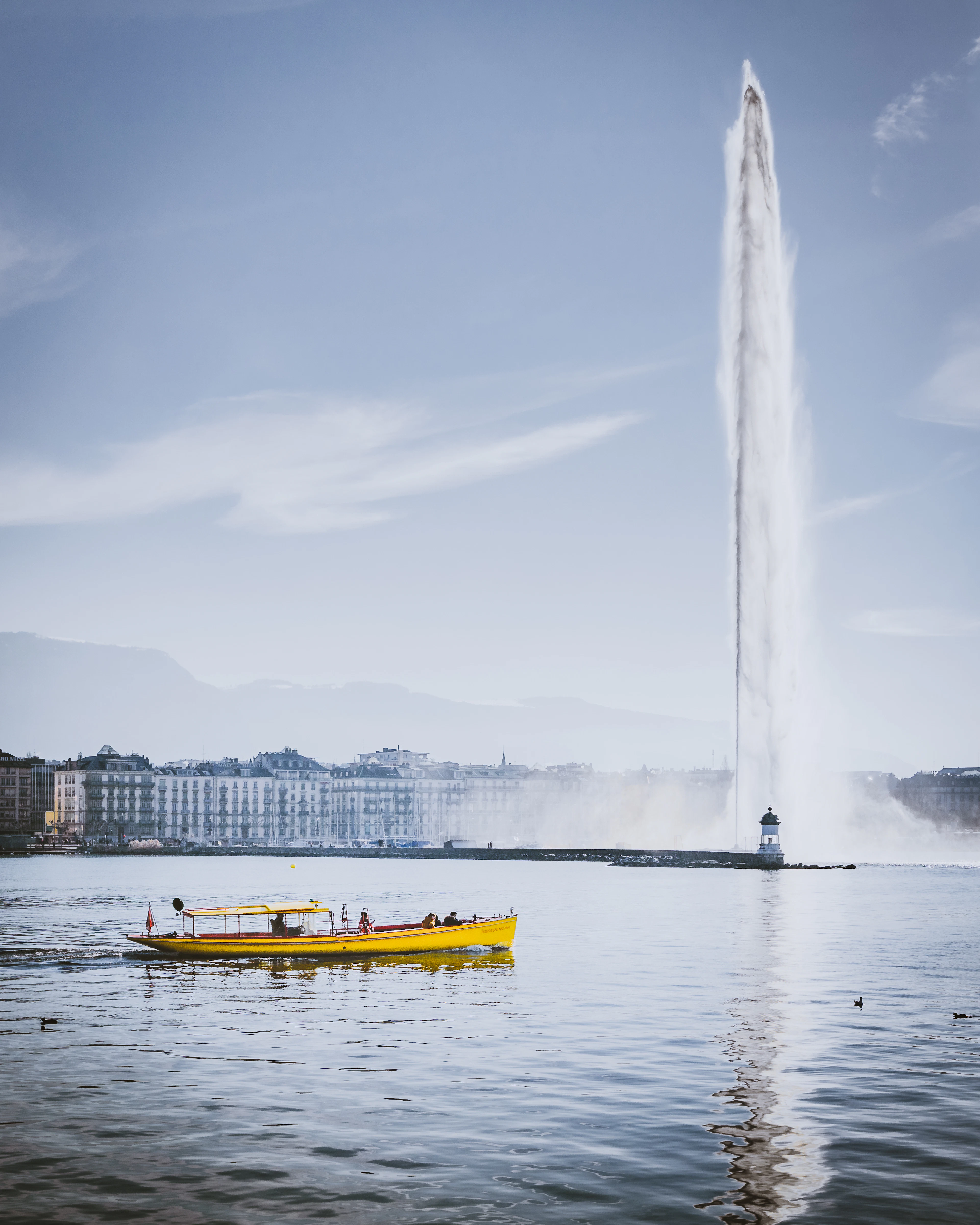 A yellow passenger boat crosses Lake Geneva in front of the towering Jet d'Eau fountain.