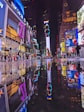 A vibrant urban nightlife scene with numerous brightly lit billboards and advertisements reflecting on a wet street surface. People are walking around, creating a lively atmosphere in a busy city square.