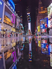 A vibrant urban nightlife scene with numerous brightly lit billboards and advertisements reflecting on a wet street surface. People are walking around, creating a lively atmosphere in a busy city square.
