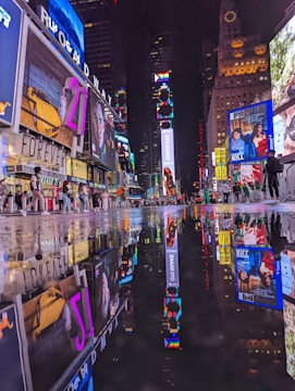 A vibrant urban nightlife scene with numerous brightly lit billboards and advertisements reflecting on a wet street surface. People are walking around, creating a lively atmosphere in a busy city square.