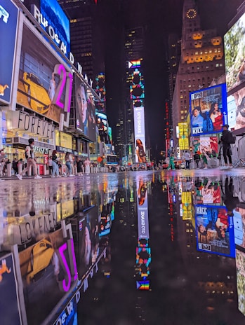 A vibrant urban nightlife scene with numerous brightly lit billboards and advertisements reflecting on a wet street surface. People are walking around, creating a lively atmosphere in a busy city square.