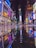 A vibrant urban nightlife scene with numerous brightly lit billboards and advertisements reflecting on a wet street surface. People are walking around, creating a lively atmosphere in a busy city square.