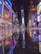 A vibrant urban nightlife scene with numerous brightly lit billboards and advertisements reflecting on a wet street surface. People are walking around, creating a lively atmosphere in a busy city square.