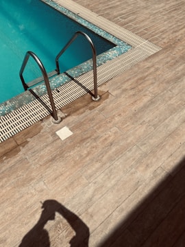 A swimming pool with turquoise water features stainless steel ladders on a wooden deck. The shadow of a person is visible on the deck, and the sunlight creates a warm and inviting atmosphere.