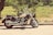 A classic cruiser motorcycle is parked on a paved road adjacent to a grassy area with lush greenery in the background. The bike features a large engine, chrome detailing, and a windshield. Sunlight casts shadows on the ground, enhancing the vintage feel of the scene.