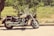 A classic cruiser motorcycle is parked on a paved road adjacent to a grassy area with lush greenery in the background. The bike features a large engine, chrome detailing, and a windshield. Sunlight casts shadows on the ground, enhancing the vintage feel of the scene.
