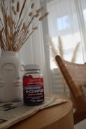 A bottle of Simply8 Gummy Vitamins, specifically B-Complex, sits on a wooden tabletop next to a white vase filled with decorative pampas grass. The scene includes a woven chair and a softly lit window with sheer curtains in the background, creating a calm and cozy atmosphere.