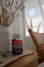 A bottle of Simply8 Gummy Vitamins, specifically B-Complex, sits on a wooden tabletop next to a white vase filled with decorative pampas grass. The scene includes a woven chair and a softly lit window with sheer curtains in the background, creating a calm and cozy atmosphere.