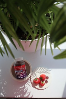 A bottle of Simply8 gummy vitamins sits in a bowl, next to a plate with three strawberries and some gummy pieces. A potted plant with green leaves partially shades the scene.