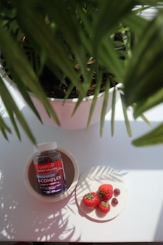 A bottle of Simply8 gummy vitamins sits in a bowl, next to a plate with three strawberries and some gummy pieces. A potted plant with green leaves partially shades the scene.