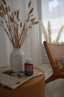 A minimalistic interior scene featuring a small round wooden table with a white ceramic vase holding dried wheat stalks. On the table, there's a bottle of B-complex vitamins and some newspapers. In the background, a woven chair and sheer white curtains partially allow light in, creating a soft and tranquil atmosphere.
