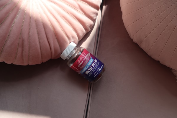 A bottle of gummy vitamins labeled 'Iron Plus', placed on a soft, neutral-toned surface between two decorative pink pillows with pleated designs.