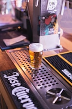 A pint of beer with a frothy head is placed on a metal drip tray at a bar counter. Behind the glass, there is a beer tap with a graphic design featuring a skull. Bar mats with distinct logos are visible on the wooden counter, adding to the pub atmosphere.