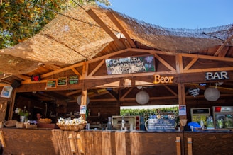 A rustic wooden bar setup under an open canopy, featuring various neon signs advertising beer and a bar. The roof is covered with a thatched material allowing sunlight to filter through, creating a warm atmosphere. Baskets with snacks, a display of drinks, and a counter with various items can be seen. A person is visible behind the bar preparing or managing the space. There are also boards with handwritten menus and decorations, suggesting a casual and welcoming environment.