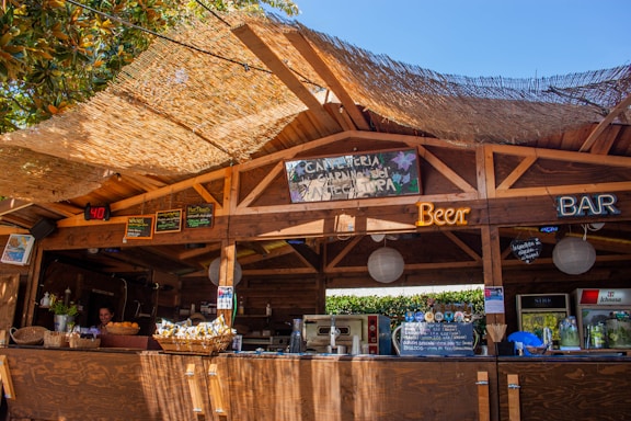 A rustic wooden bar setup under an open canopy, featuring various neon signs advertising beer and a bar. The roof is covered with a thatched material allowing sunlight to filter through, creating a warm atmosphere. Baskets with snacks, a display of drinks, and a counter with various items can be seen. A person is visible behind the bar preparing or managing the space. There are also boards with handwritten menus and decorations, suggesting a casual and welcoming environment.