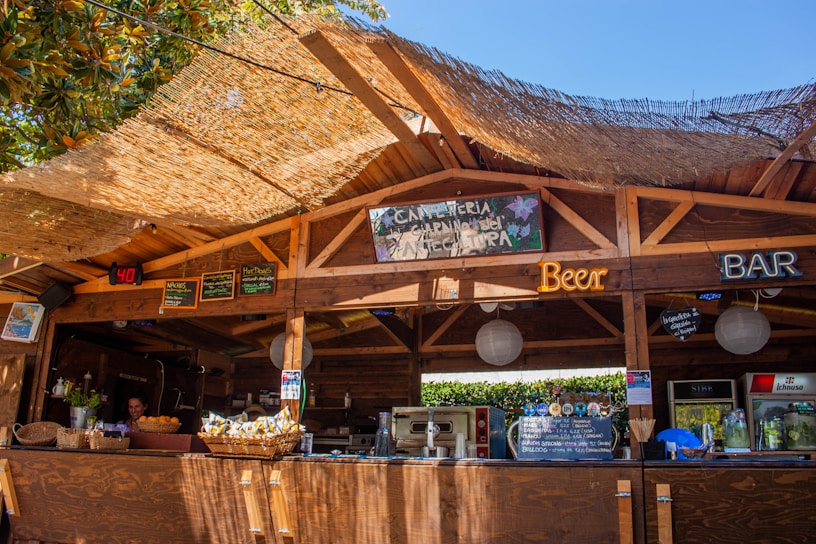 A rustic wooden bar setup under an open canopy, featuring various neon signs advertising beer and a bar. The roof is covered with a thatched material allowing sunlight to filter through, creating a warm atmosphere. Baskets with snacks, a display of drinks, and a counter with various items can be seen. A person is visible behind the bar preparing or managing the space. There are also boards with handwritten menus and decorations, suggesting a casual and welcoming environment.