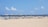 A sandy beach features rows of lounge chairs arranged under a series of blue and white striped canopy structures. There are people in the background, some under umbrellas, and the ocean can be seen with a clear blue sky and a few clouds above.