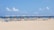 A sandy beach features rows of lounge chairs arranged under a series of blue and white striped canopy structures. There are people in the background, some under umbrellas, and the ocean can be seen with a clear blue sky and a few clouds above.