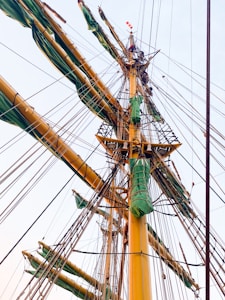 Tall sailing ship masts with multiple ropes and rigging, featuring green covers on the yardarms. The structure appears complex and robust against a clear sky, highlighting the intricate rigging and sails.