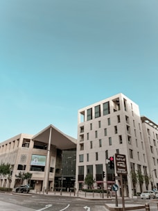 A modern architectural structure with clean lines and a mix of glass and stone elements is present. The building has multiple stories, featuring large windows and balconies. There is a prominent entrance to the Galleria, with trees and a clear blue sky in the background. Vehicles are present on the street, and there are traffic signs, including one in both English and Arabic indicating to look right for the tramway.